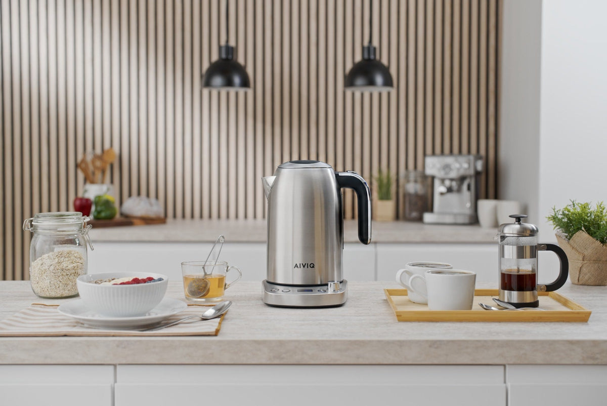 Wasserkocher aus Edelstahl auf einer Arbeitsplatte mit Frühstück, Tee in einem Glas und einer French Press mit zwei Tassen auf einem Tablett