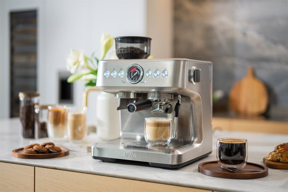 Espressomaschine aus Edelstahl mit Bohnenbehälter und Manometer, ein Glas Kaffee mit Milch unter dem Auslauf und ein schwarzer Kaffee auf einem Holzbrett daneben