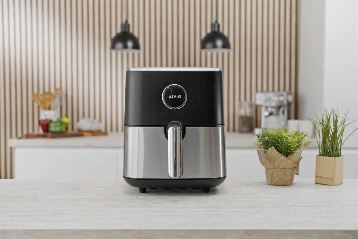 Schwarzer Airfryer mit Schublade aus Edelstahl auf einer hellen Arbeitsplatte zwischen zwei grünen Pflanzen, mit einer Espressomaschine im Hintergrund