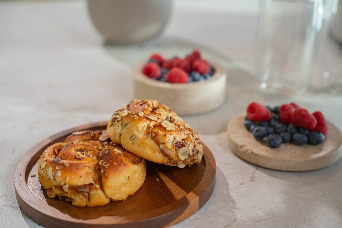 Schnecken mit Mandeln auf einer runden Holzplatte, mit zwei Schalen Himbeeren und Blaubeeren und einem Glas im Hintergrund