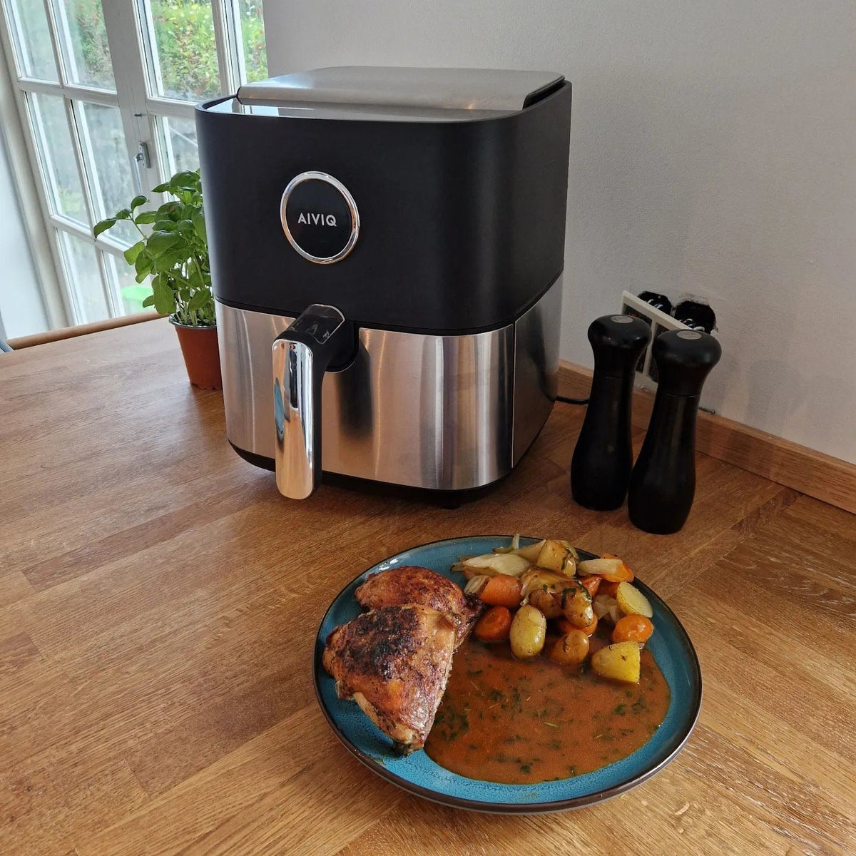 Premio Airfryer auf einem Holztisch neben einem Teller mit Hähnchenschenkeln, Kartoffeln und Soße