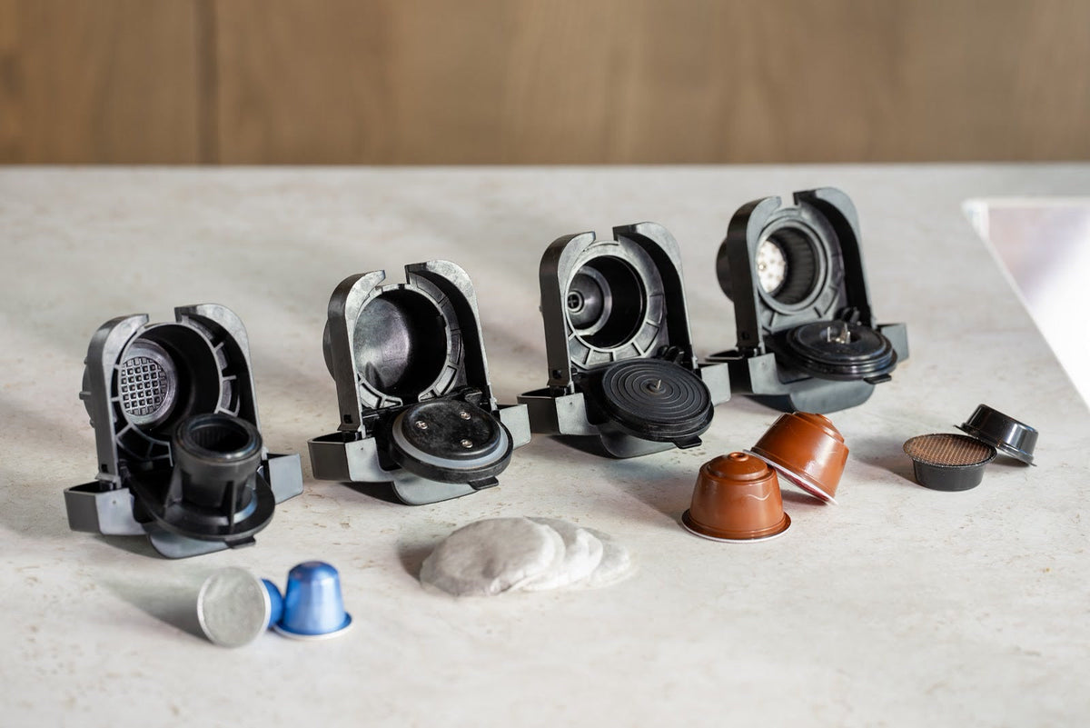 Vier geöffnete Kapseladapter auf einem Tisch mit Kapseln in verschiedenen Formen und weichen Kaffeepads