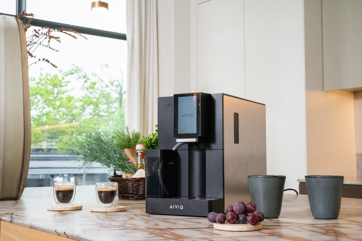 Schwarze Espressomaschine mit Touchscreen und Milchbehälter auf einem Marmortisch am Fenster, mit zwei Espressi in Gläsern, zwei Bechern und Weintrauben