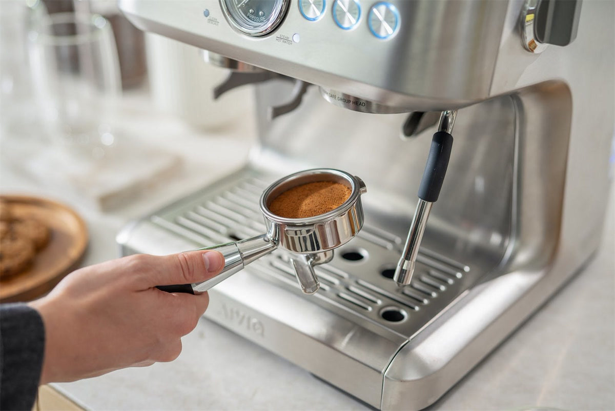 Eine Hand hält einen Siebträger mit getampertem Kaffee vor einer Espressomaschine aus Edelstahl