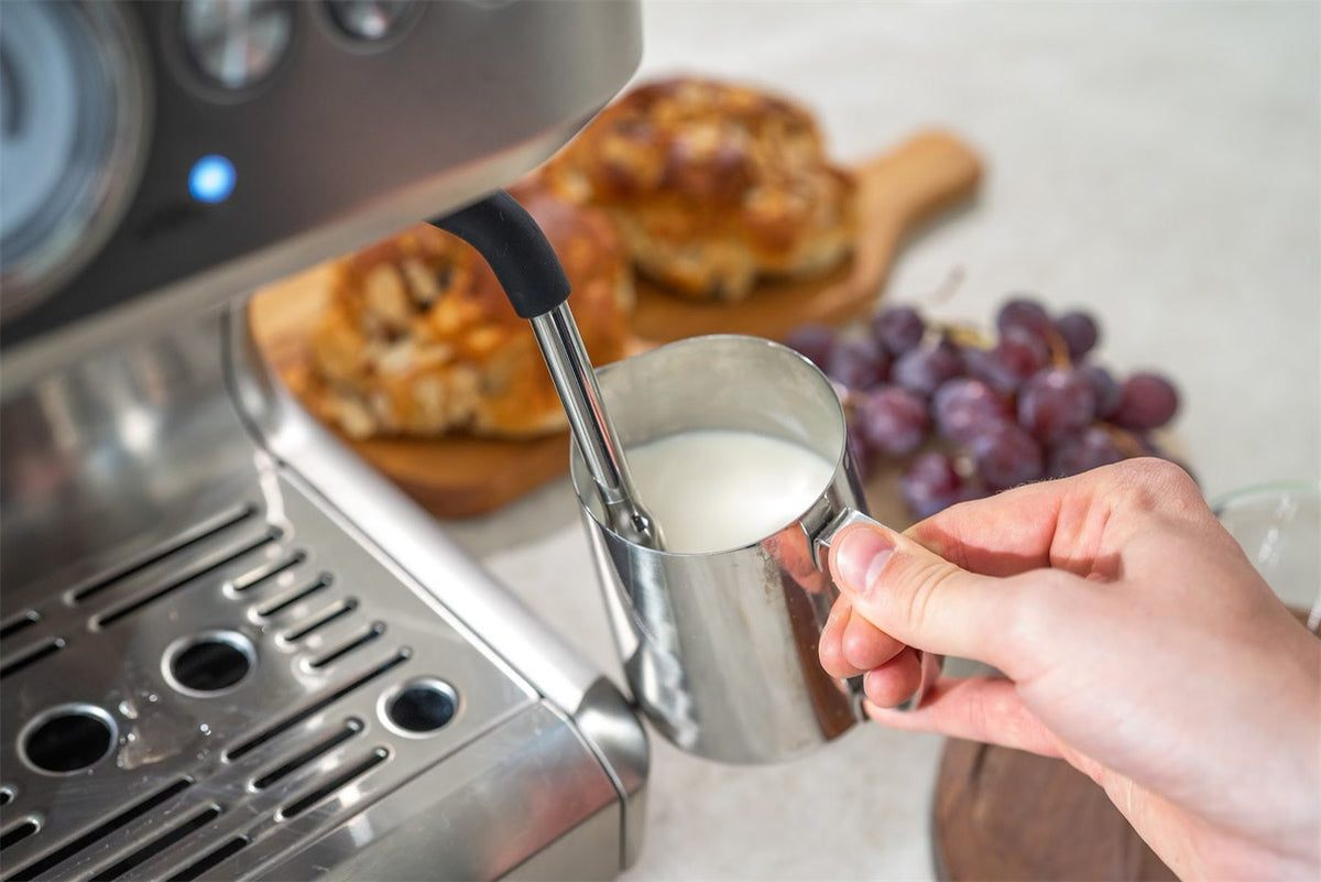 Eine Hand hält ein Edelstahlkännchen mit Milch unter die Dampflanze einer Espressomaschine aus Edelstahl, dahinter Brötchen und Weintrauben