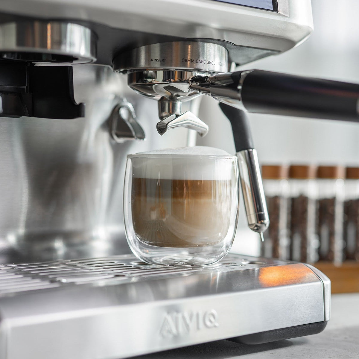Ein doppelwandiges Glas mit Kaffee, Milch und Schaum in Schichten steht auf der Abtropfschale unter dem Brühkopf. Rechts hängt die Dampflanze, die einen Temperaturfühler hat.