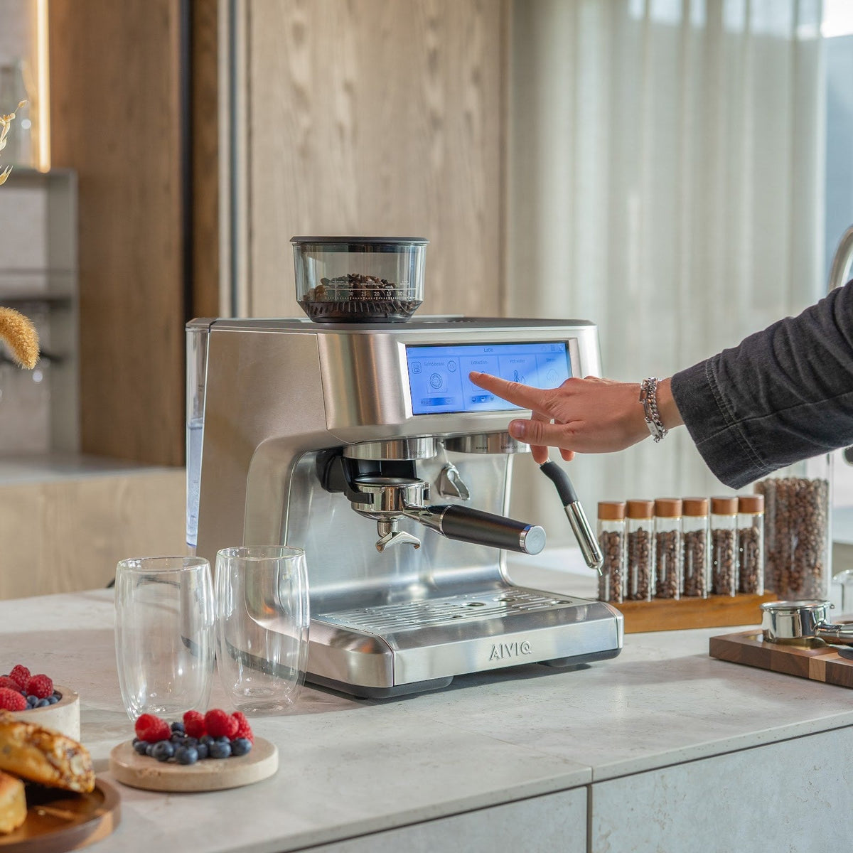 Eine Person tippt in einer hellen Küche auf den Touchscreen der Aspire Expert. Auf der Arbeitsplatte stehen Gläser, Beeren und Kaffeebohnen in Glasröhrchen.