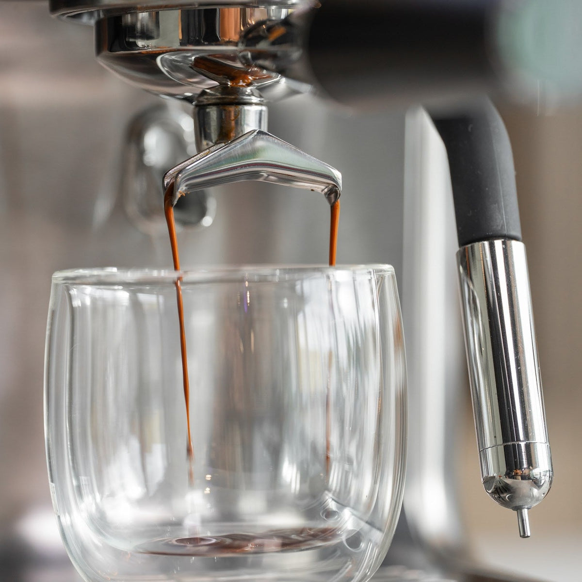 Espresso läuft in zwei dünnen Strahlen aus dem Auslauf des Siebträgers in ein doppelwandiges Glas. Rechts hängt die Dampflanze.